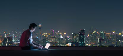 A person using a laptop on a rooftop at night with a city skyline in the background